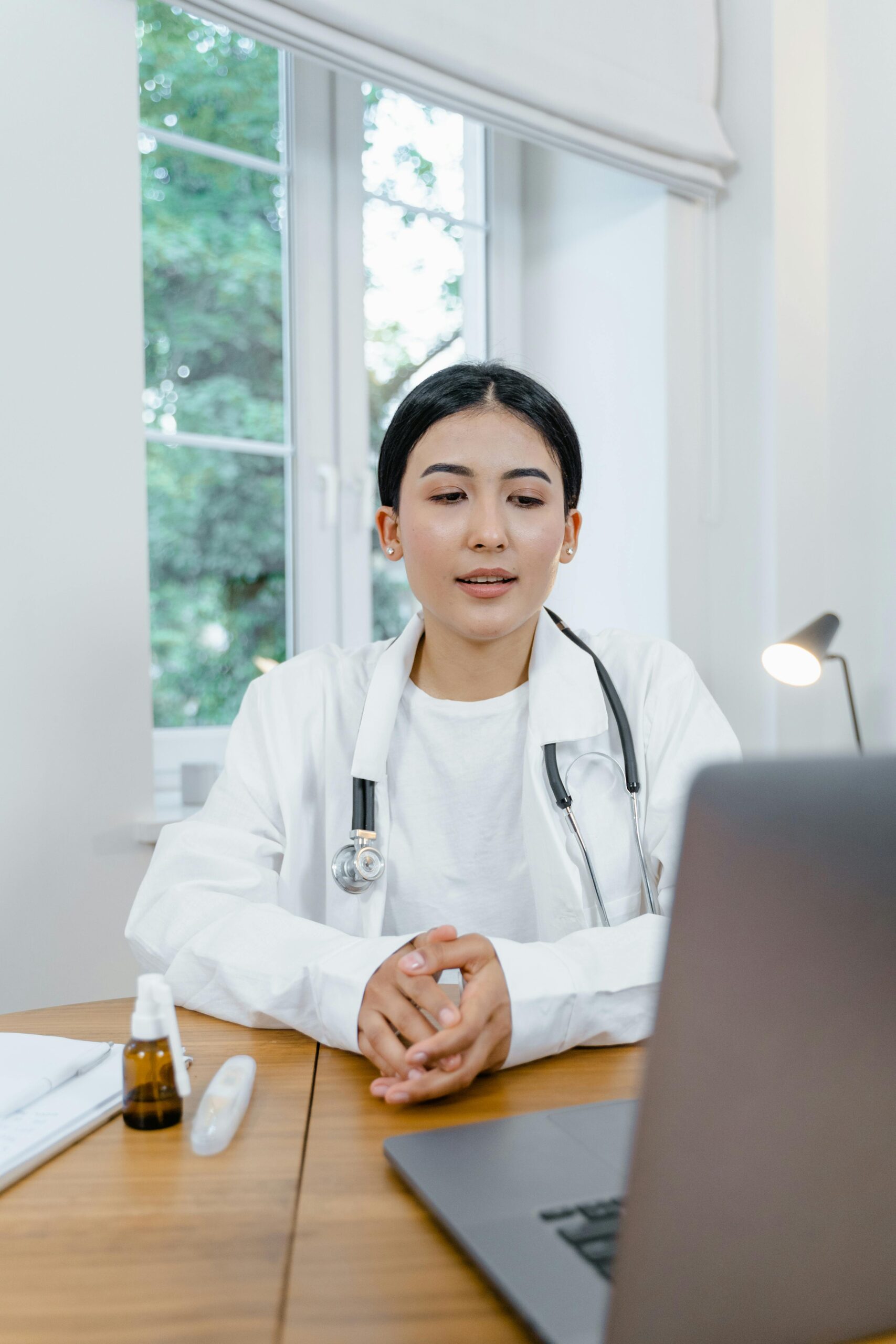 Asian female doctor in white coat using a laptop for online consultation from her office desk.